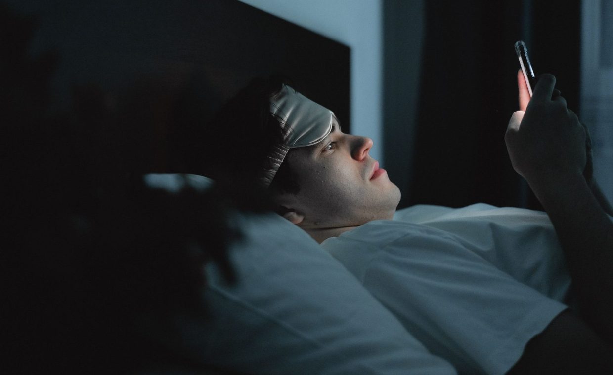 A man wearing a sleep mask late at night using a smartphone on his bed.