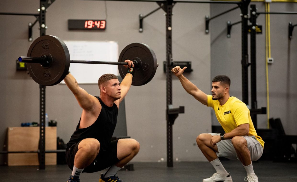 Man lifting weights under coach supervision in a gym setting, focusing on strength training.