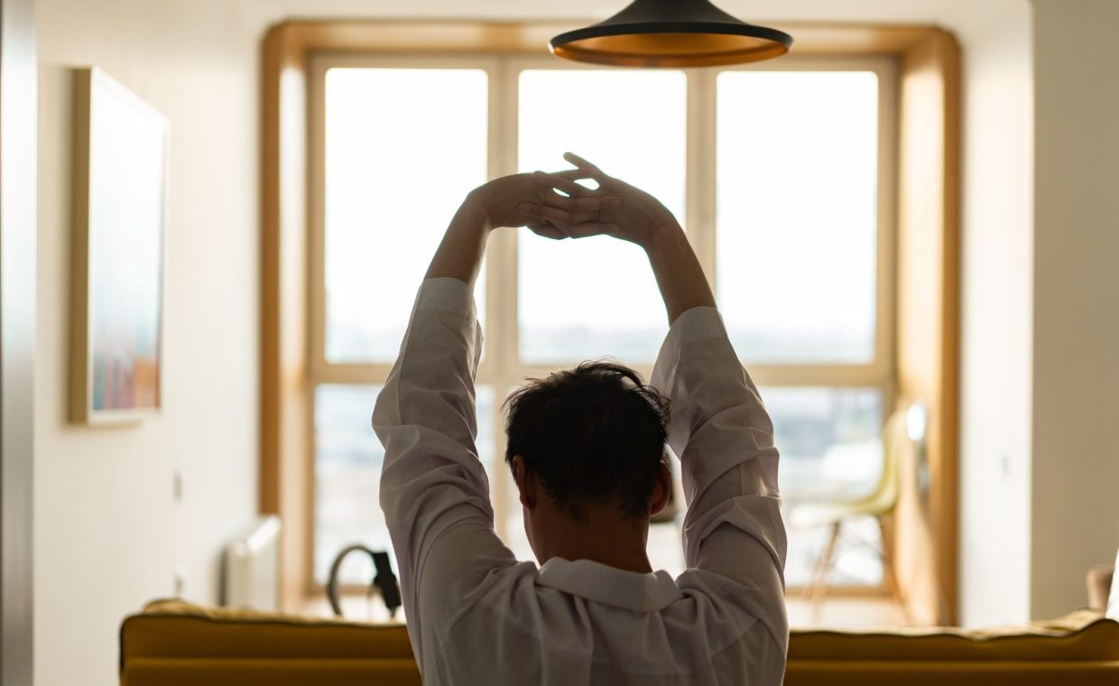Back view of a man stretching inside a room with a sunny window backdrop.