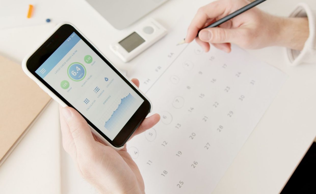 Close-up of hands using a smartphone to track health stats while planning on a calendar.