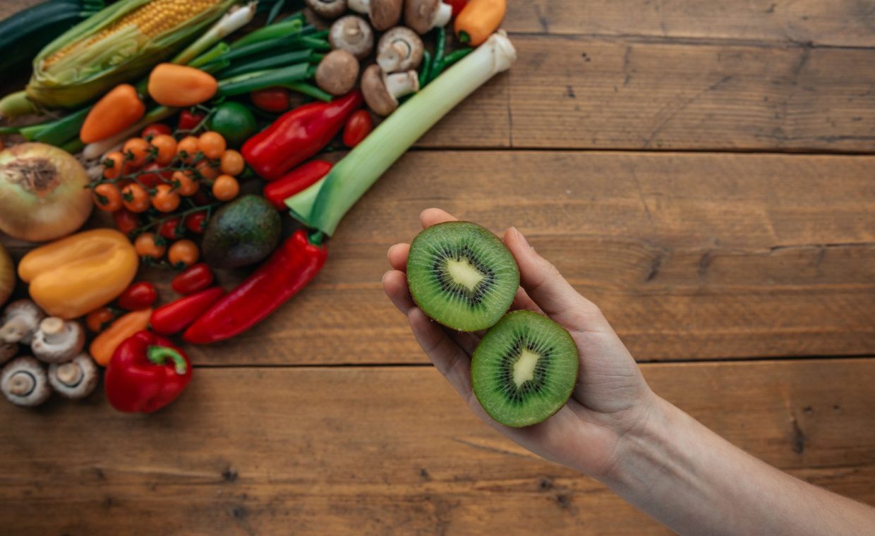 A hand holds sliced kiwi above a wooden table with fresh vegetables, highlighting healthy ingredients.