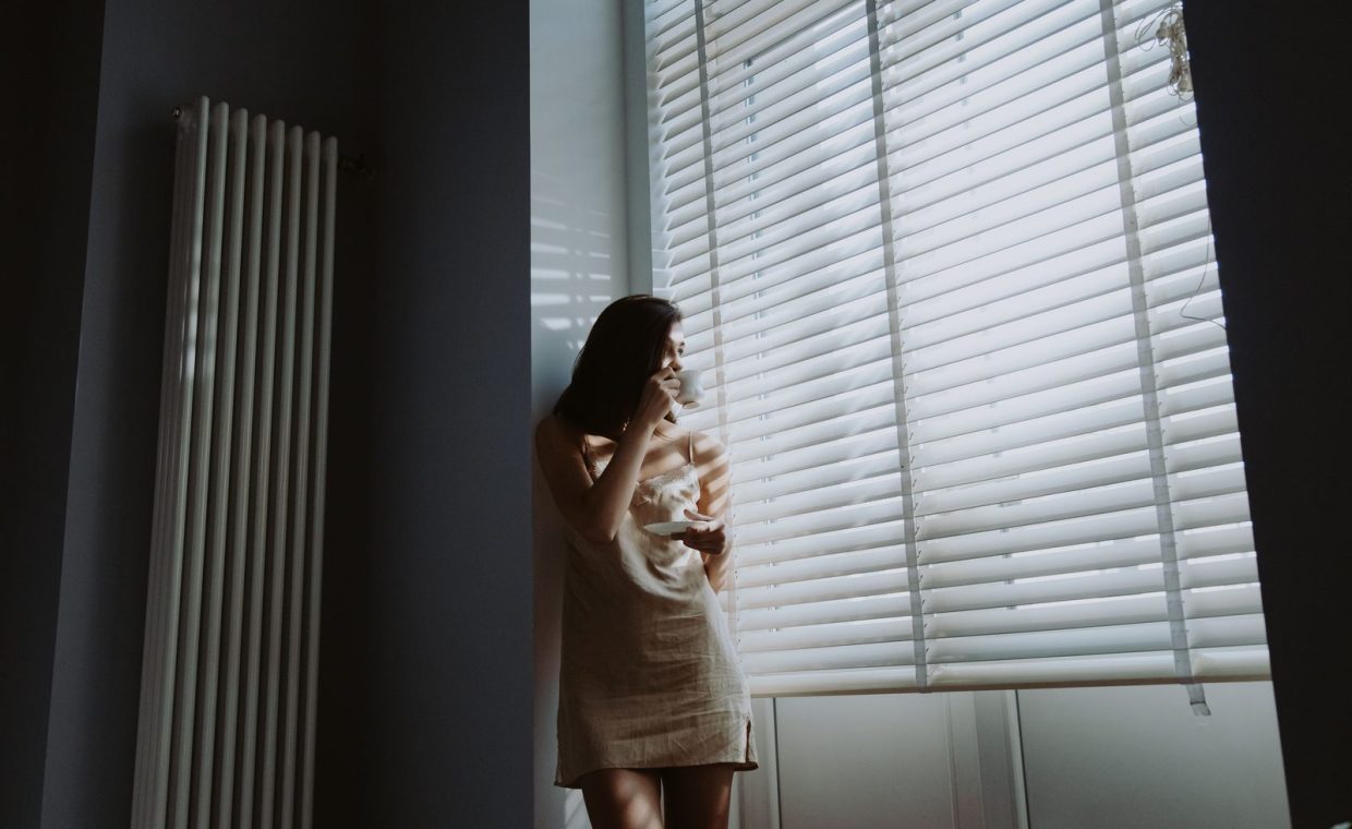 A woman in soft morning light stands by the window, holding a cup and saucer, savoring a peaceful moment.