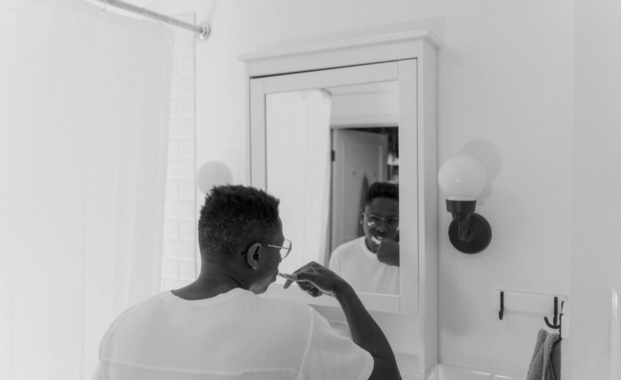 A black and white photo of a man brushing his teeth in front of a bathroom mirror.