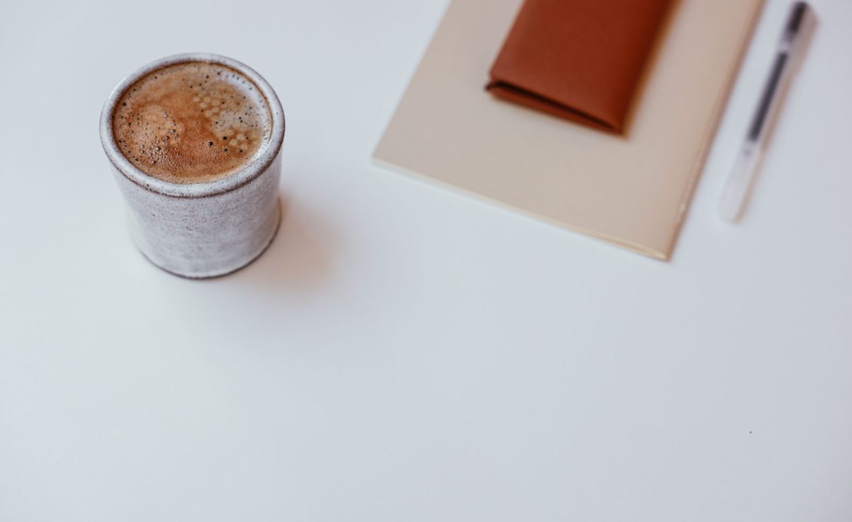 A minimalist setup with a cup of coffee, a notebook, and a pen on a white tabletop.