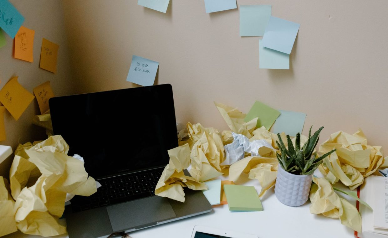 A chaotic office desk with a laptop, scattered papers, sticky notes, and a potted plant.