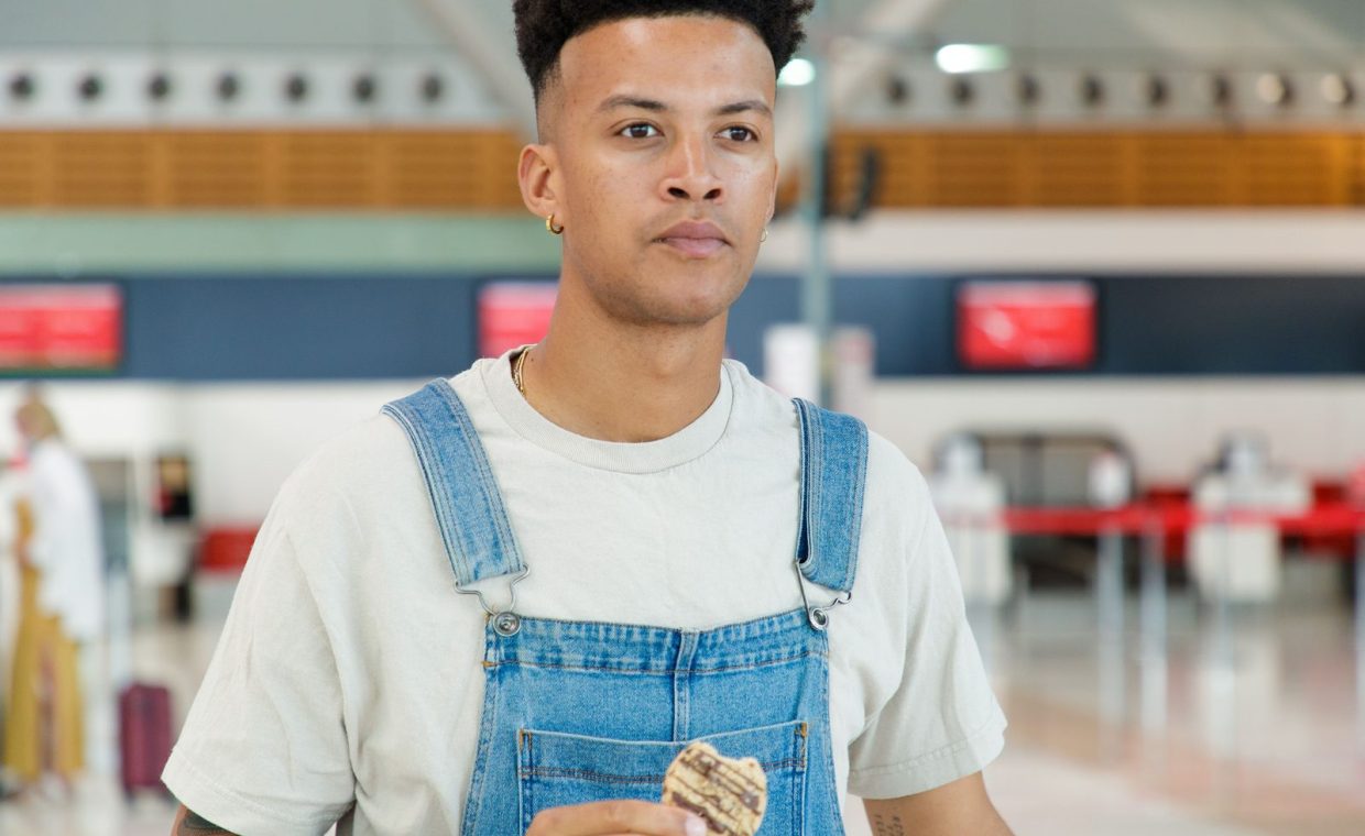 Casual young man in denim overalls eating a cookie at an airport terminal.