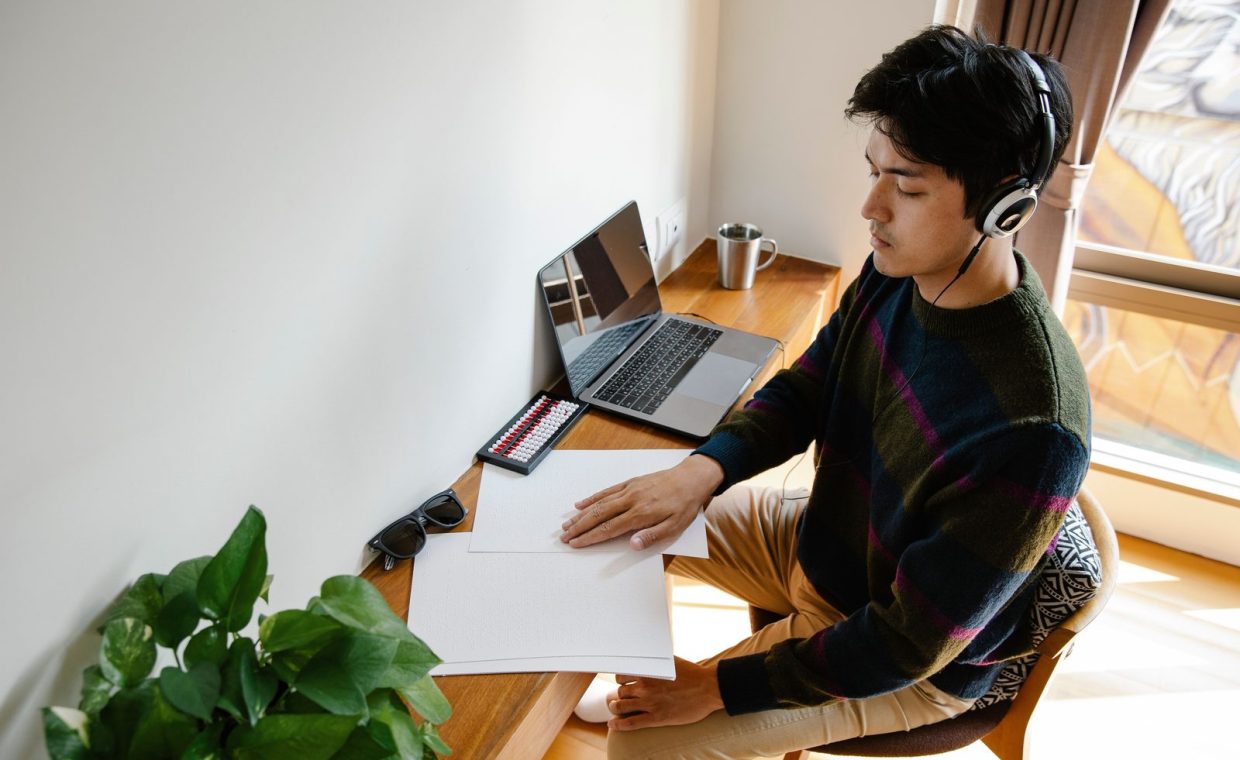 A man wearing headphones concentrates while working at a desk with plants and a laptop.