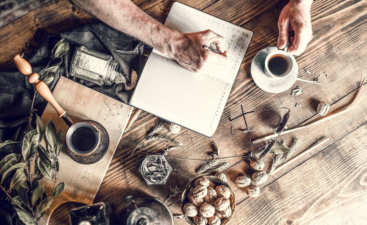 From above senior person taking notes while having coffee at wooden table with scattered walnuts and plants with jezve