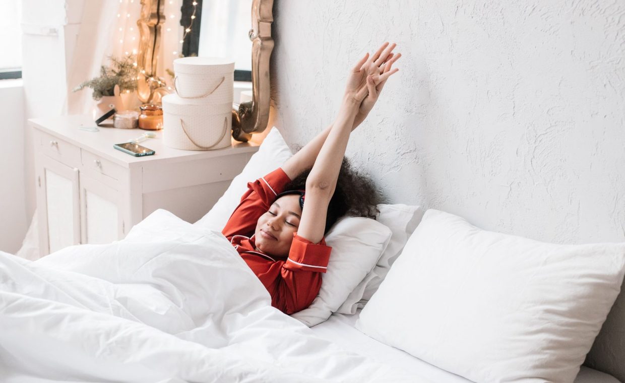 Woman in red pajamas stretching in a cozy, minimalist bedroom. Bright morning light enhances the serene scene.