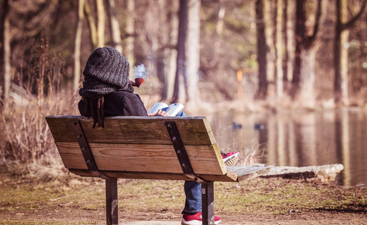 A person relaxes on a wooden bench by a lake, reading a book and exhaling smoke.