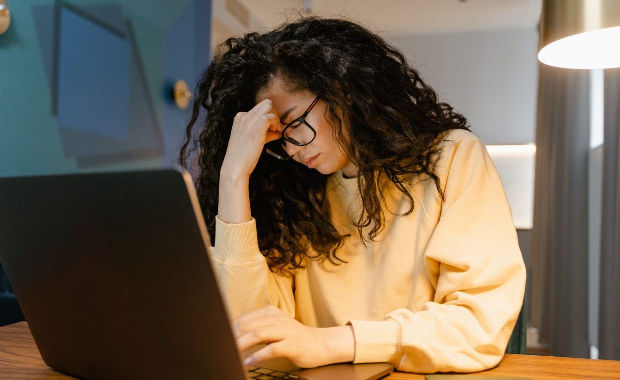 A tired woman with curly hair and glasses experiencing stress while working on a laptop.