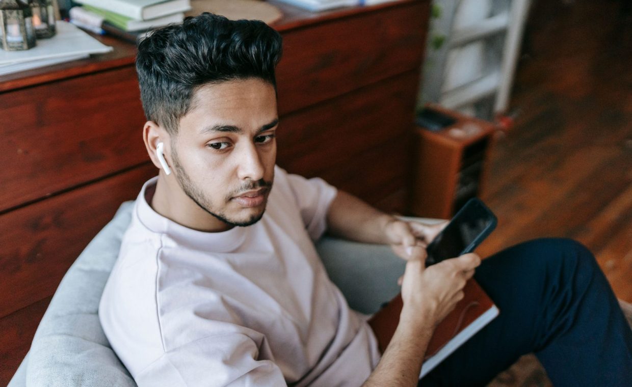 Dreamy young ethnic male in casual clothes browsing mobile phone and listening to music via earbuds while sitting in arm