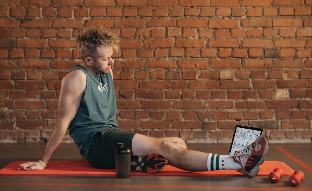 Athletic man sitting and resting on a mat in an urban gym, with a tablet and weights visible.