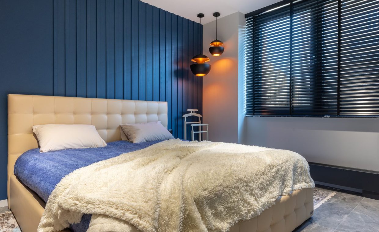 Modern bedroom interior with blanket and pillows on bed against lamps and jalousie in house