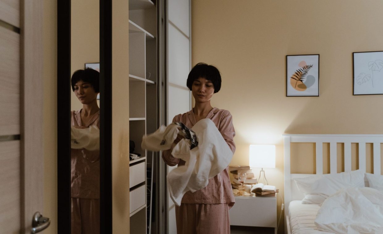 A woman in pajamas tidying up her closet in a warmly lit bedroom at night.