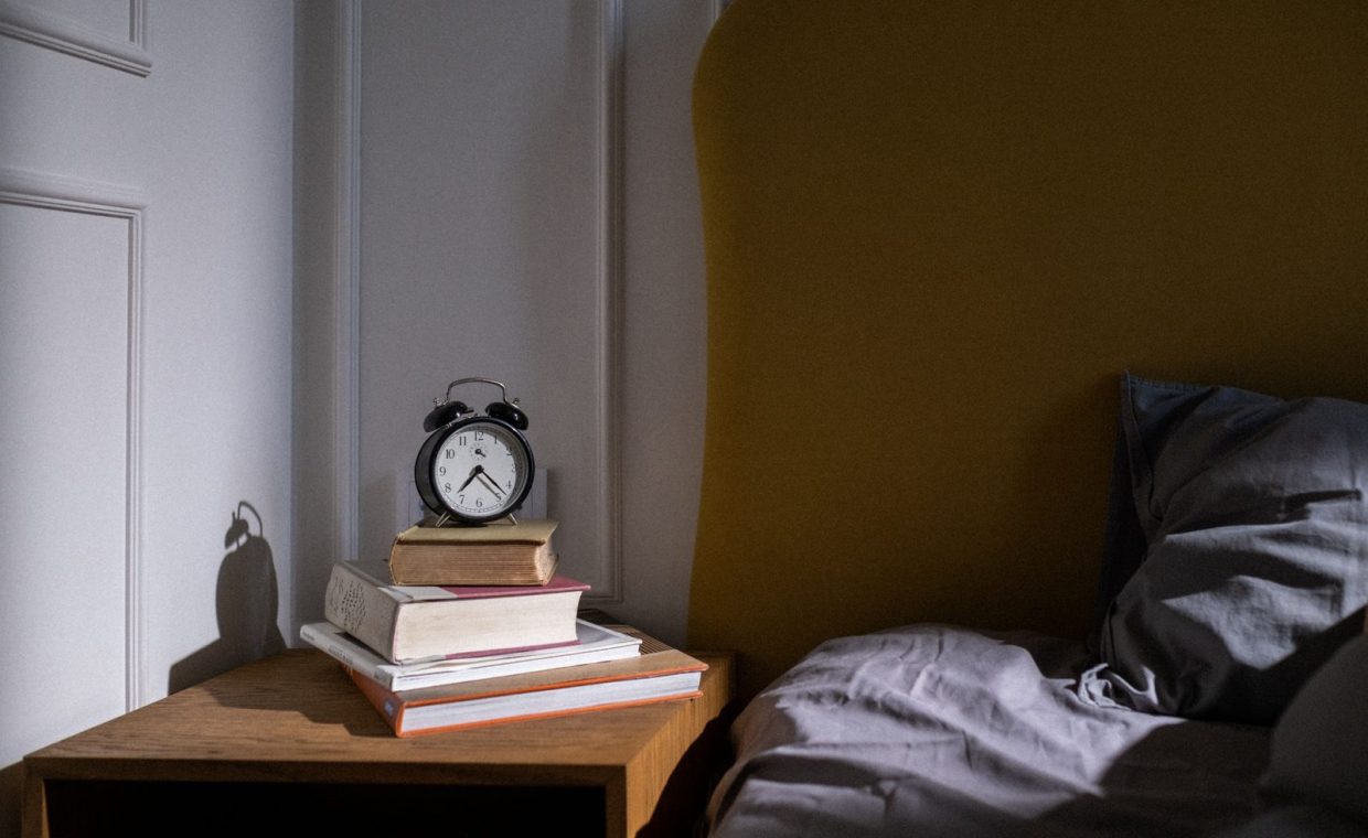 A cozy bedroom setting with books and an alarm clock on a nightstand next to a bed.