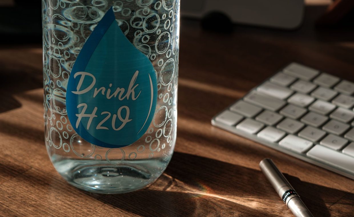 Glass water bottle labeled 'Drink H2O' on an office desk with sunlight and keyboard.