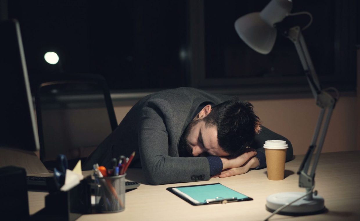 Man in office suit sleeping on desk with coffee and tablet nearby, symbolizing work fatigue.
