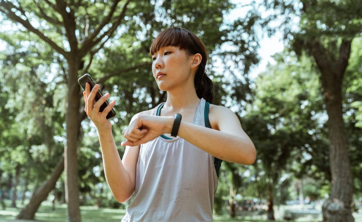 Positive concentrated young fit Asian sportswoman in activewear browsing smartphone and checking fitness bracelet while