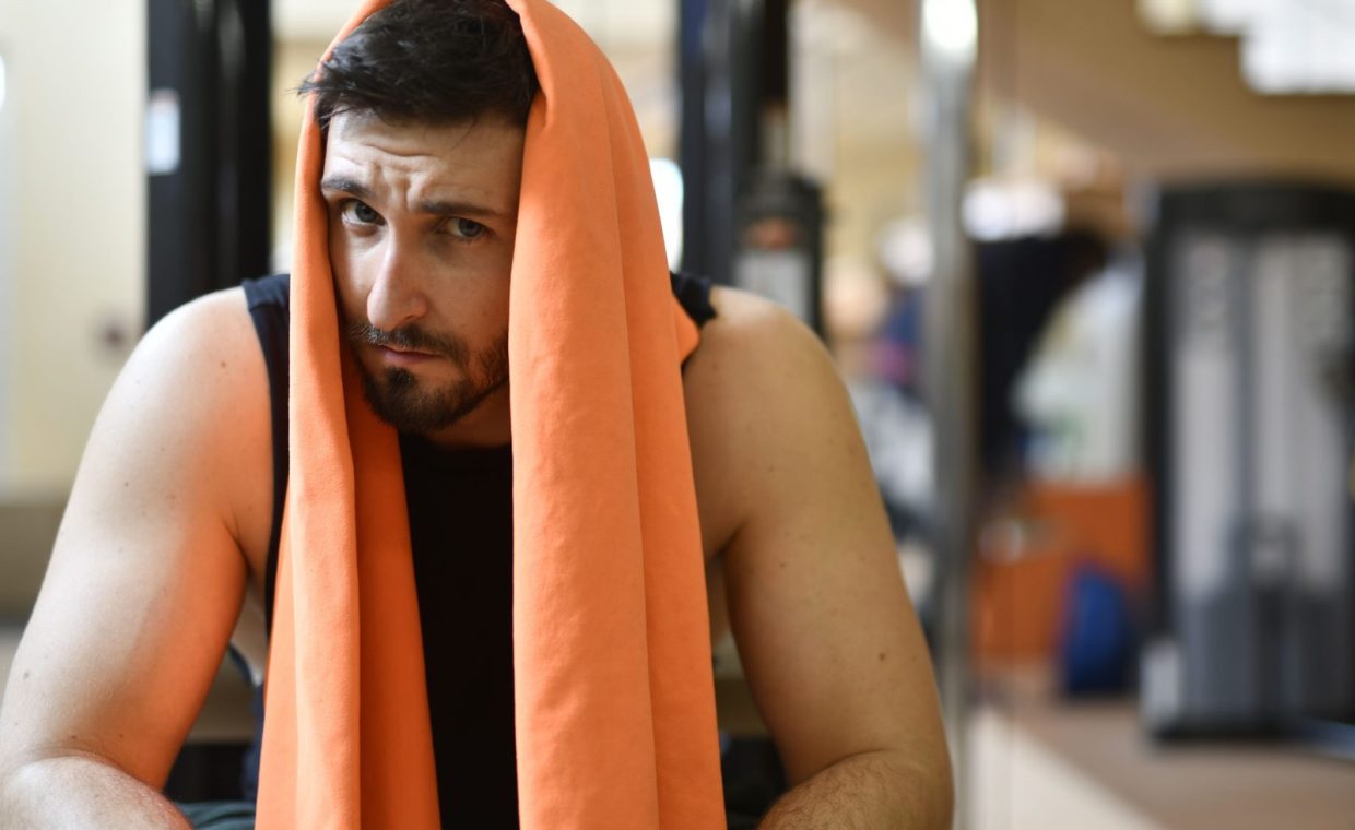 A tired man rests with an orange towel after a workout in the gym.